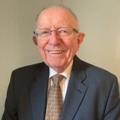 Michael, an older, light skinned man with receding grey hair and glasses smiles happily at the camera. He's wearing a grey suit, white shirt and beige patterned tie.