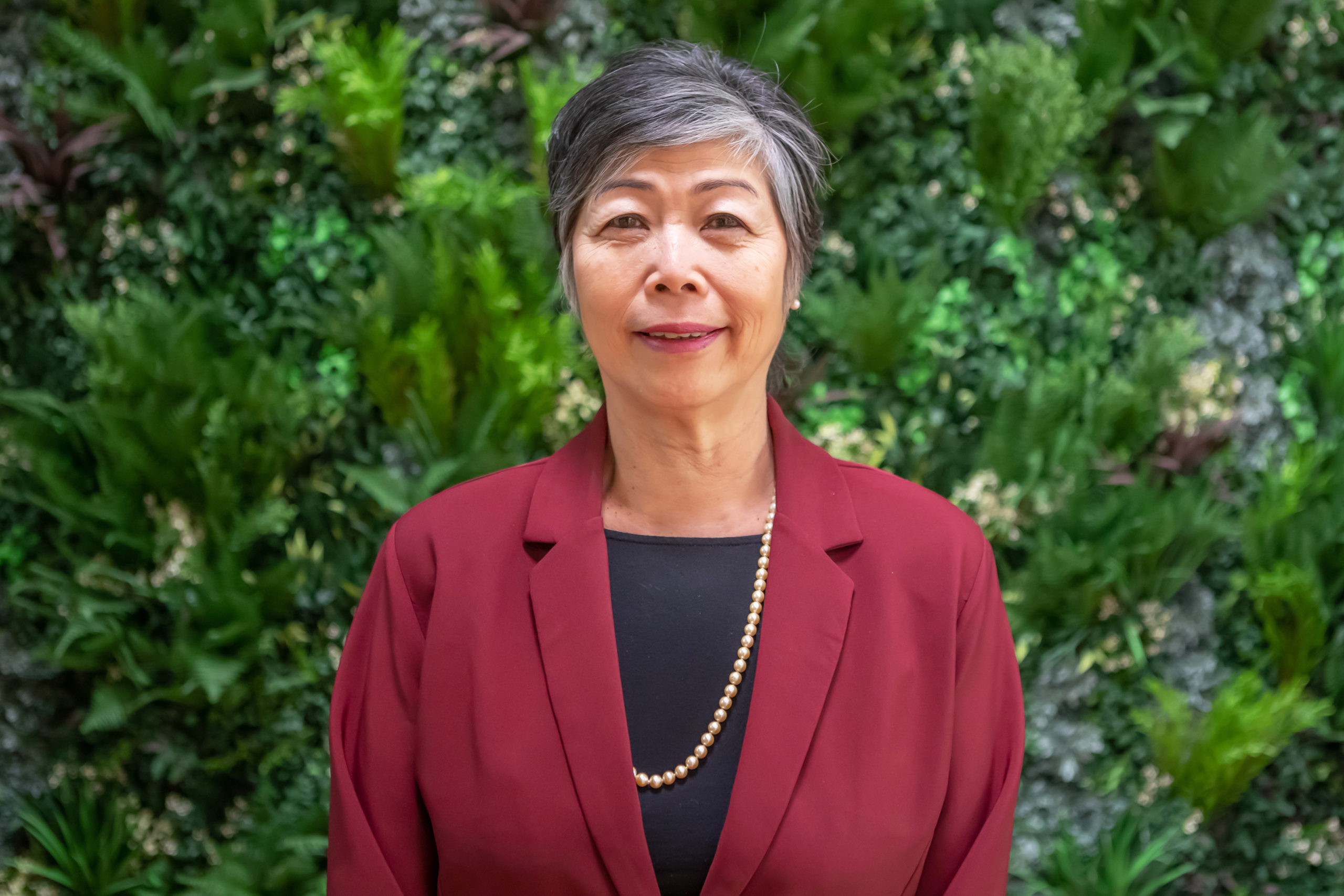 Eileen, a medium-light skinned woman with short grey hair stands in front of a wall of lush green foliage. She's smiling, and wears a burgundy coloured jacket, black top and long gold necklace.
