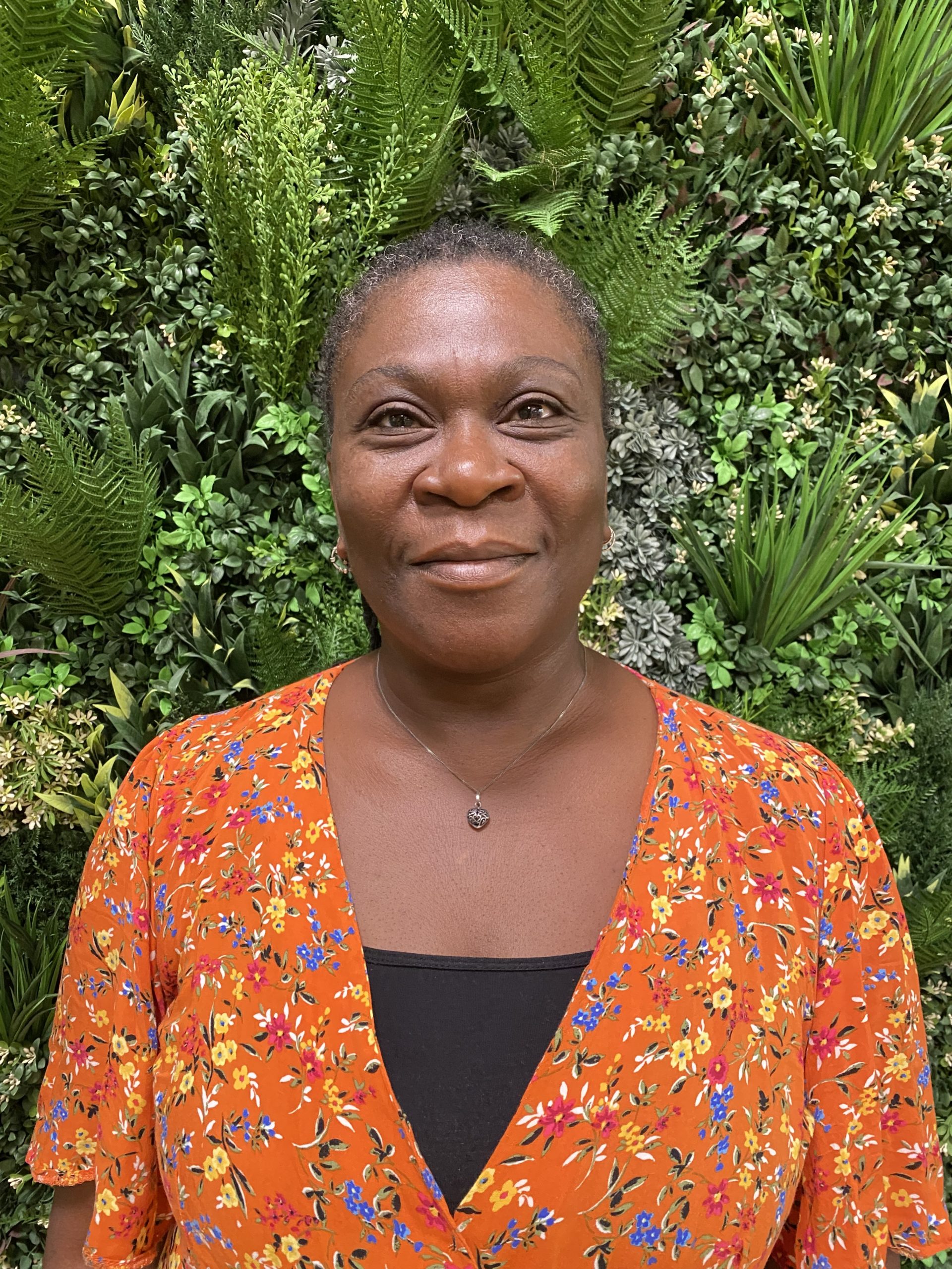Bee, a dark skinned woman with dark hair flecked with grey smiles as she stands in front of a wall of lush green foliage. She is wearing a bright orange patterned top with a black top underneath.