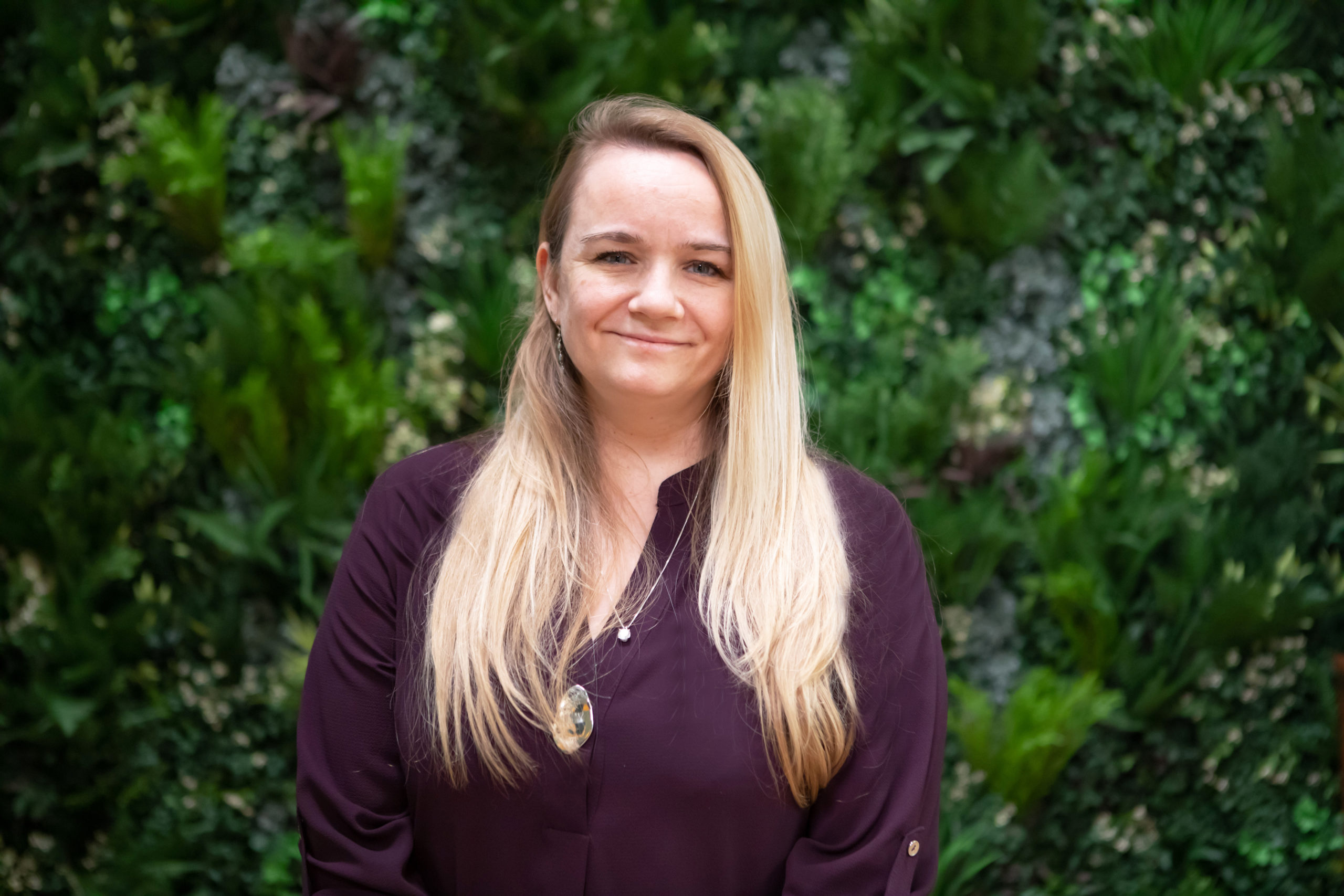 Josie, a light skinned woman with long blonde hair is smiling and looking straight ahead. She's standing against a wall covered in green foliage, and wears a purple top.