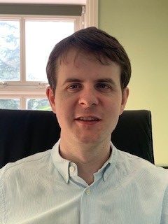 Callum, a light skinned man with dark hair and wearing a white open necked shirt faces the camera directly, giving a half-smile.