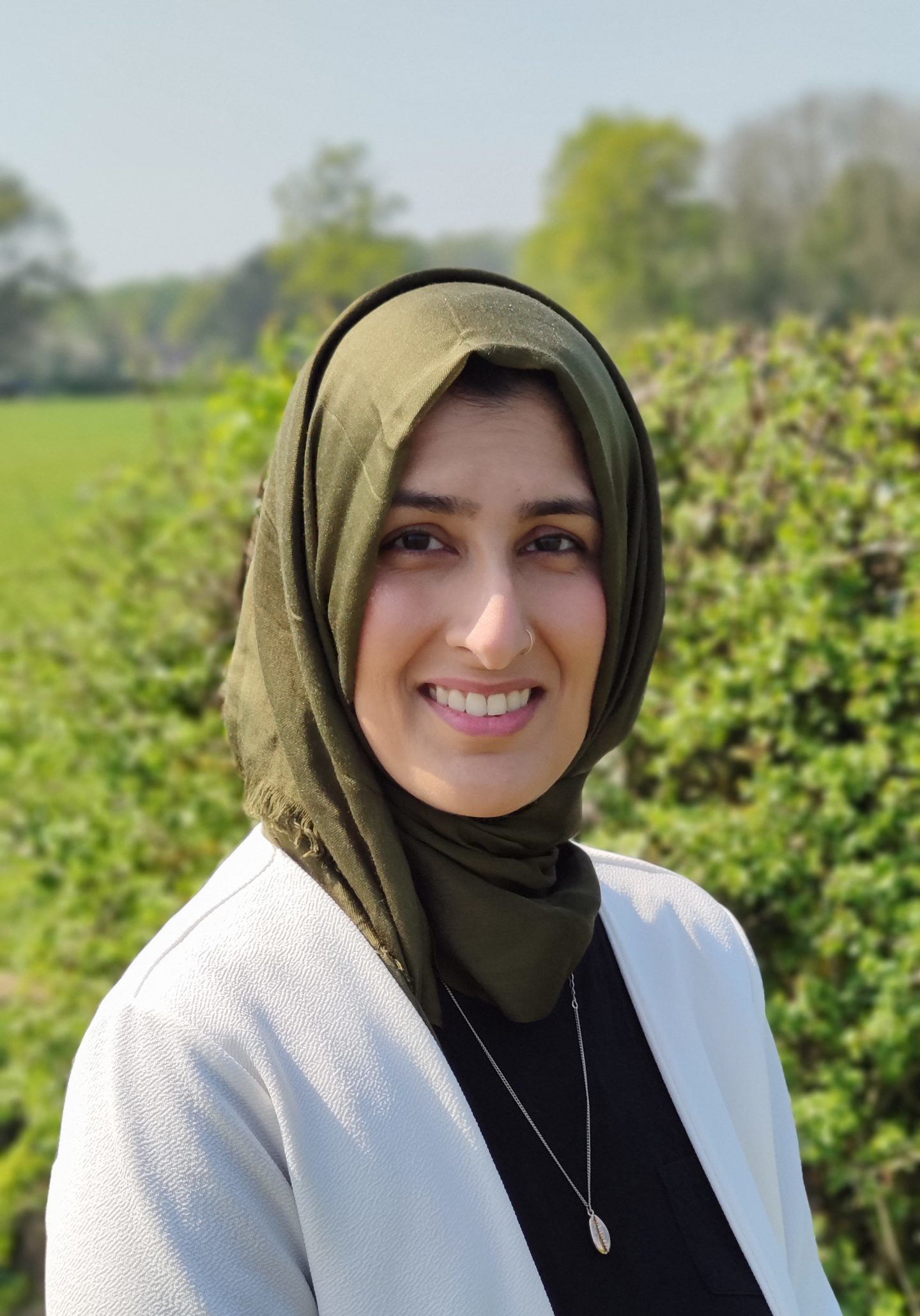 Sabira, a medium light skinned woman looks directly at the camera. She is in a field with vegetation in the background and is wearing a white cardigan with a black top underneath, a long necklace and a headscarf.