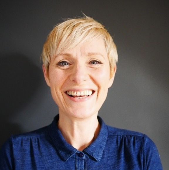 Helen, a light skinned woman with short blonde hair wearing a blue shirt stands in front of a dark background and laughs into the camera.