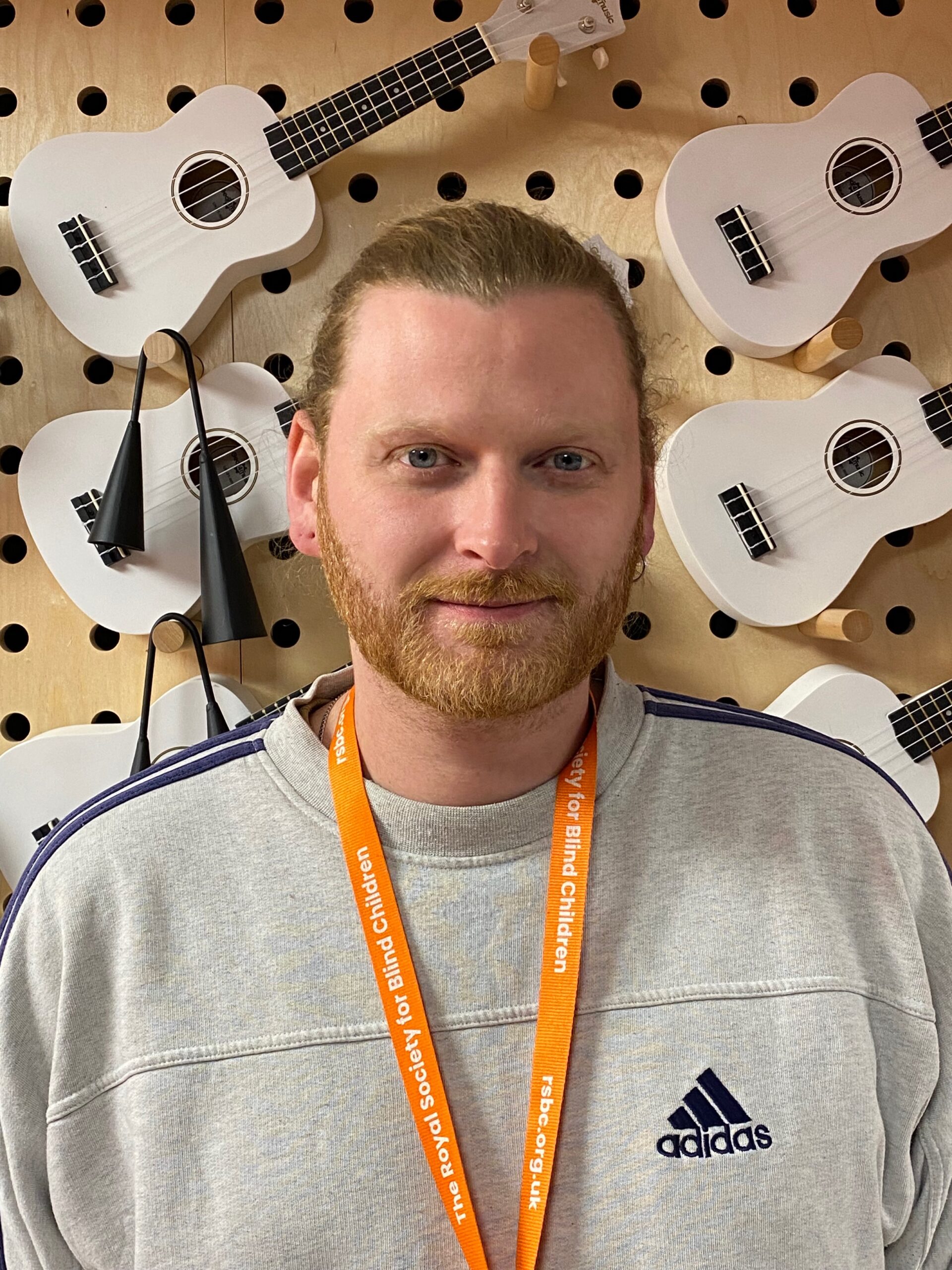 Owain, a light skinned man with red hair and a beard, wears an RSBC lanyard and a grey sweatshirt and smiles as he looks into the camera.