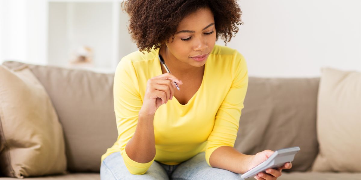 A medium skinned woman with short, curly brown hair wears a yell top and sits on a beige sofa looking at her calculator, holding a pen.