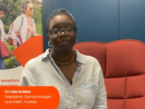 A woman sitting on a red chair, in front of an RSBC banner. The text reads:"Dr Lola Solebo, Paediatric Ophtalmologist and RSBC Trustee"