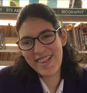 Camille, a young person with black rimmed glasses and long dark hair, wears a black jacket and white shirt and smiles broadly at the camera.