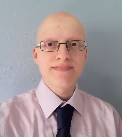 Ali, a light skinned man with glasses, wears a light coloured shirt with a dark tie and has a slight smile as he looks straight ahead at the camera.