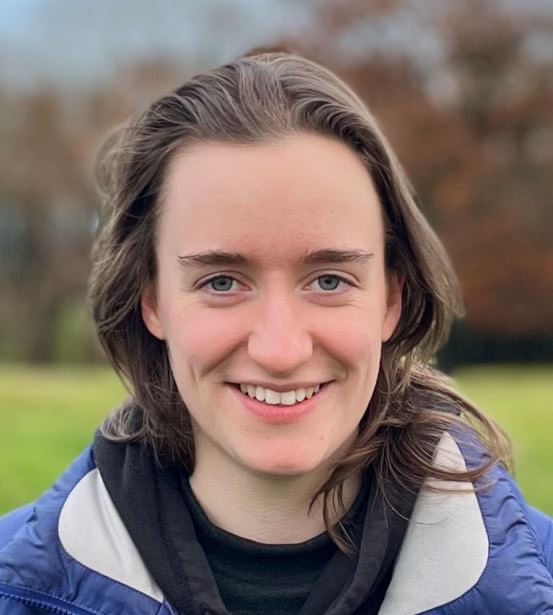 Fran, a light skinned woman with shoulder length dark hair, smiles broadly into the camera. She wears a blue padded jacket and a black top.