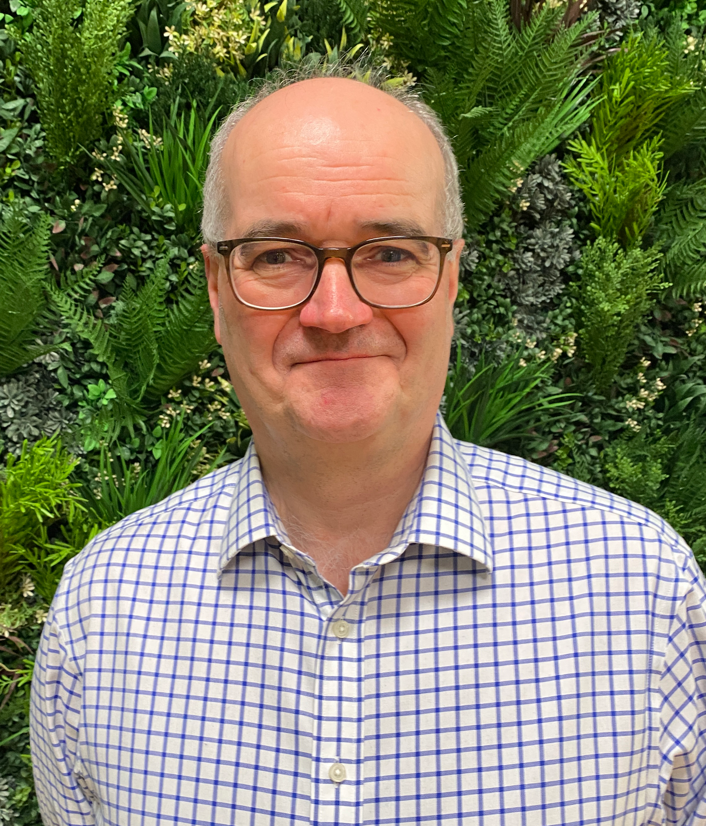 Tom, a light skinned man with grey hair and glasses wears a white shirt with blue checks and half-smiles into the camera. He is standing in front of a wall covered in green vegetation.