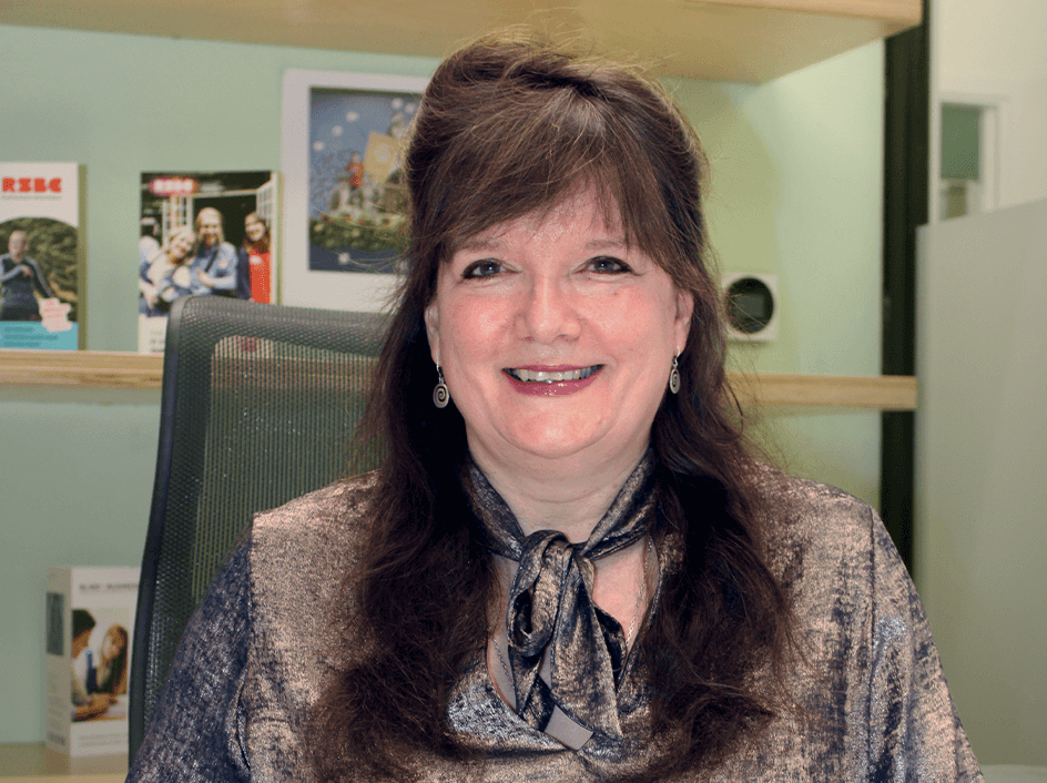 Julie, a light skinned woman with long brown hair sits at a desk with her hands folded in front of her. There are wooden shelves in the background. She is smiling broadly and wearing a metallic bronze coloured top.