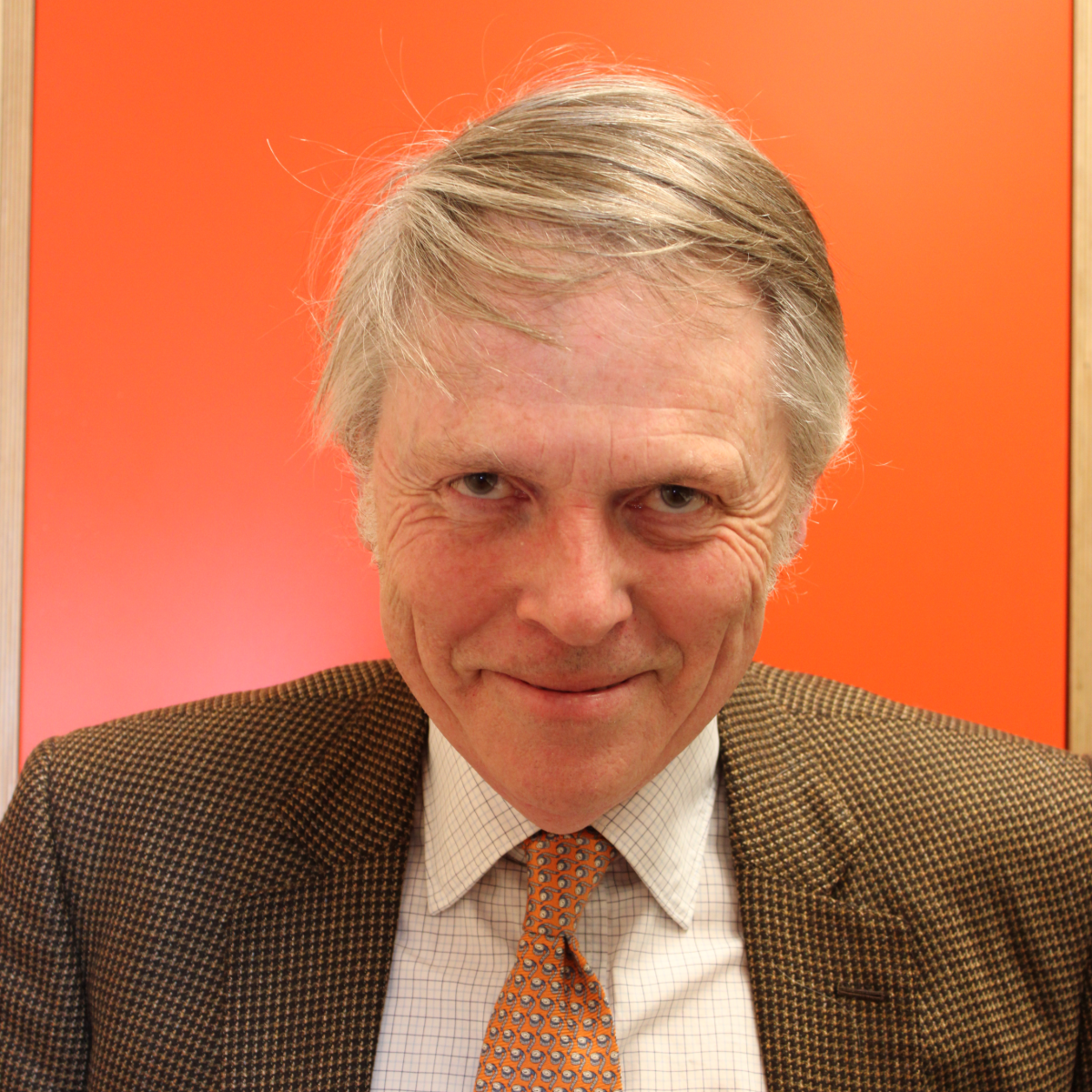 William, a light skinned man with grey hair wears a brown suit with an orange checked tie and stands in front of an orange background, smiling at the camera.