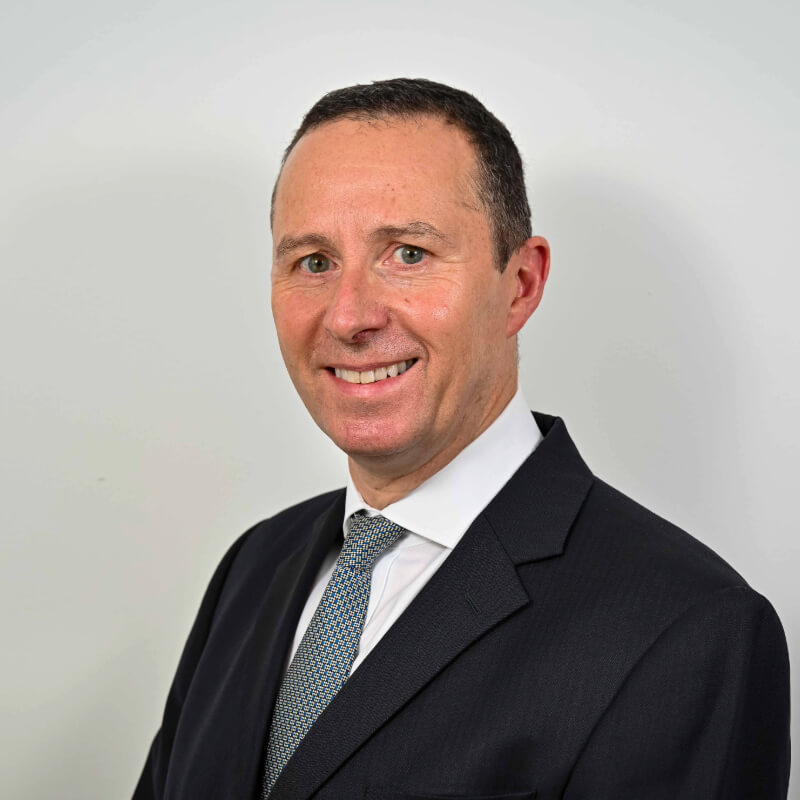 Peter, a light skinned man with dark hair in a dark suit, white shirt and grey tie sits side-on and smiles brightly at the camera.