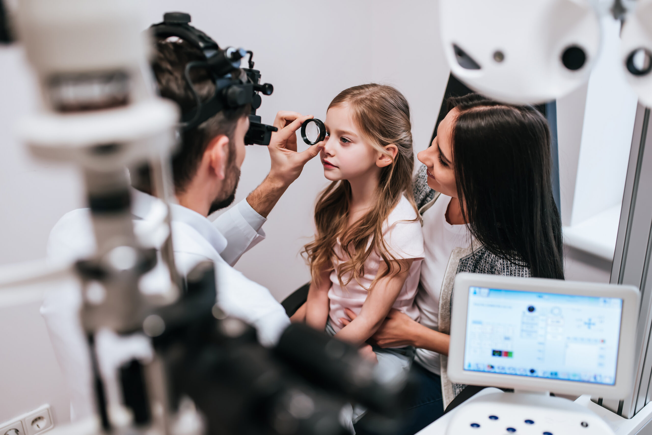 A light skinned mother sits holding her young daughter and looking at her as a light skinned optician with a beard carries out an eye test in a clinic. There is clinical technology in the foreground.
