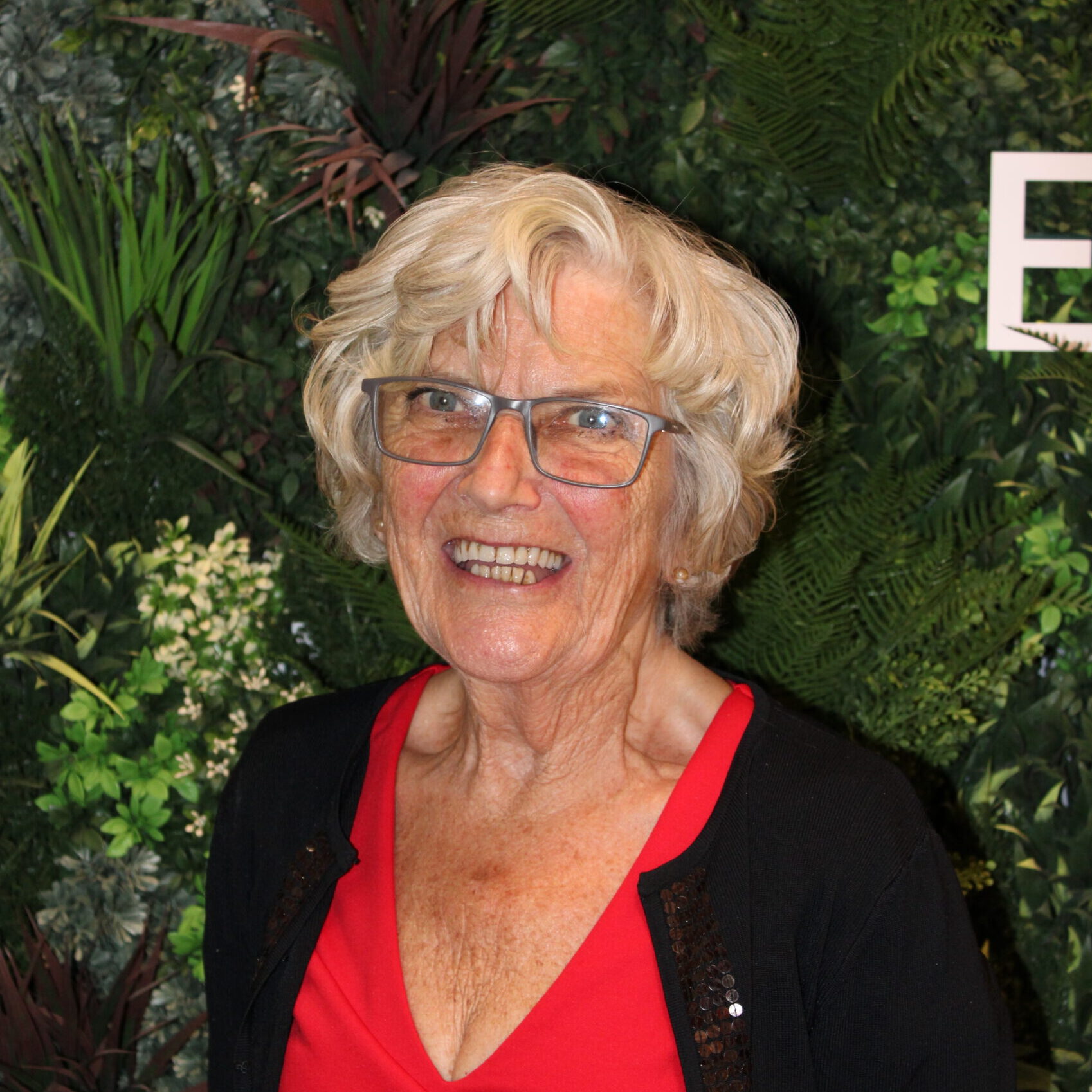 Headshot of Val May, an RSBC Trustee. Val is a light-skinned woman with white hair in a short bob style. She is wearing a red shirt with a black cardigan and titanium grey glasses. She is smiling brightly at the camera. Behind her is a wall of green foliage.