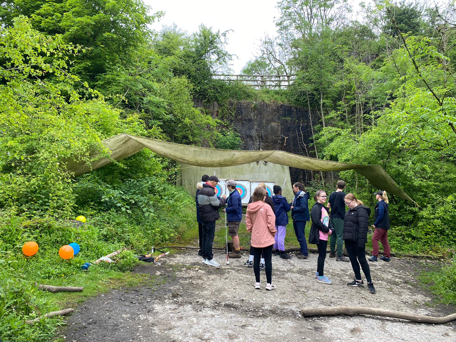 A group of young people at an RSBC residential trip. Outside, in a green wooden area, teh young people are gathered in front of an archery base. Behind them are archery targets and an instructer preparing people to begin archery.