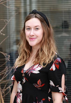 Amy has wavy auburn hair and is smiling. She wears a black headband and dark a floral top.