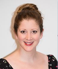 Ellie is smiling. She has brown hair styled in a high bun, fair skin and wears red lipstick and a dark top with a floral pattern.