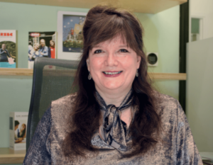 Julie, a light skinned woman with long brown hair sits at a desk with her hands folded in front of her. There are wooden shelves in the background. She is smiling broadly and wearing a metallic bronze coloured top.