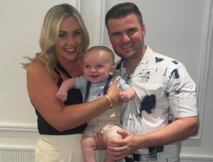 A light-skinned mother and father, Abbie and Lewis, holding their young boy, Albie. They are all smiling brightly. Abbie is wearing a black and white dress, Lewis, a white shirt with black spots on it and Albie a blue shirt with cream dungarees.