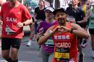 Ricardo, a 2025 RSBC London Marathon runner wearing an RSBC orange running vest with RSBC's logo, his name and running number: 3180 on. He is holding his hands up making a heart sign with them and looking directly at the camera. In the background there is a diverse number of runners in a variety of coloured running vests running for other charities.