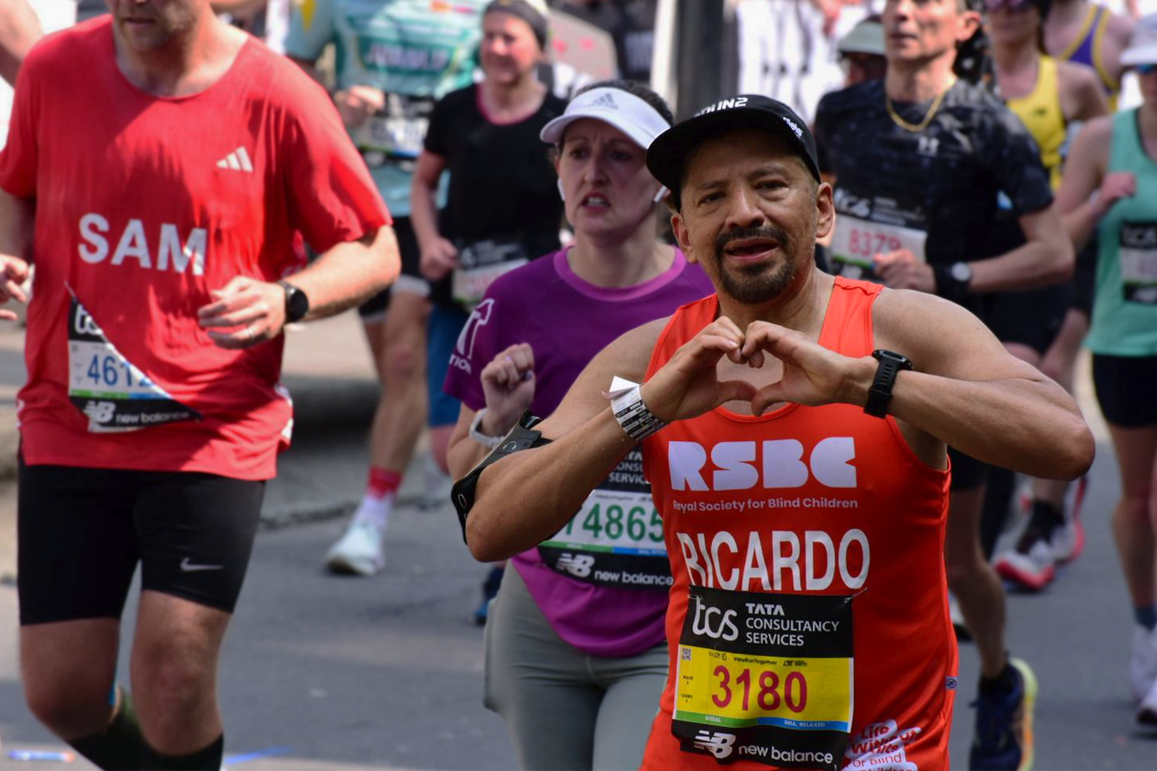 Ricardo, a 2025 RSBC London Marathon runner wearing an RSBC orange running vest with RSBC's logo, his name and running number: 3180 on. He is holding his hands up making a heart sign with them and looking directly at the camera. In the background there is a diverse number of runners in a variety of coloured running vests running for other charities.