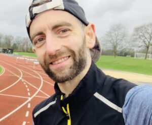 Nick, a light-skinned man who's a London Marathon fundraiser for RSBC taking a selfie. He's smiling at the camera wearing a long-sleeved blue top, a black sleeveless running jacket over the top and a black cap.