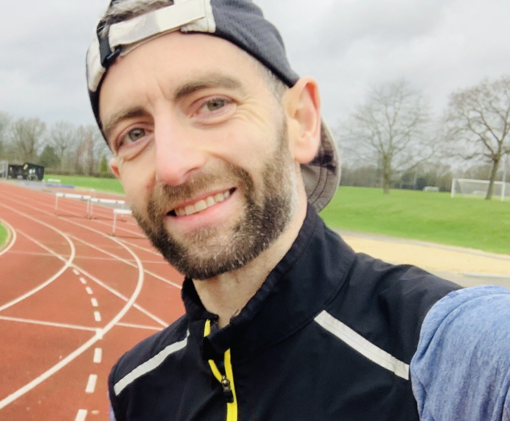 Nick, a light-skinned man who's a London Marathon fundraiser for RSBC taking a selfie. He's smiling at the camera wearing a long-sleeved blue top, a black sleeveless running jacket over the top and a black cap.
