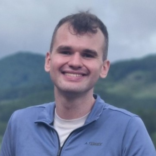 Travis, a light-skinned male wearing a blue top with a v-neck zip and a white high-neck top underneath. He is smiling to the camera and there is a lush green forest and countryside hills in the background.