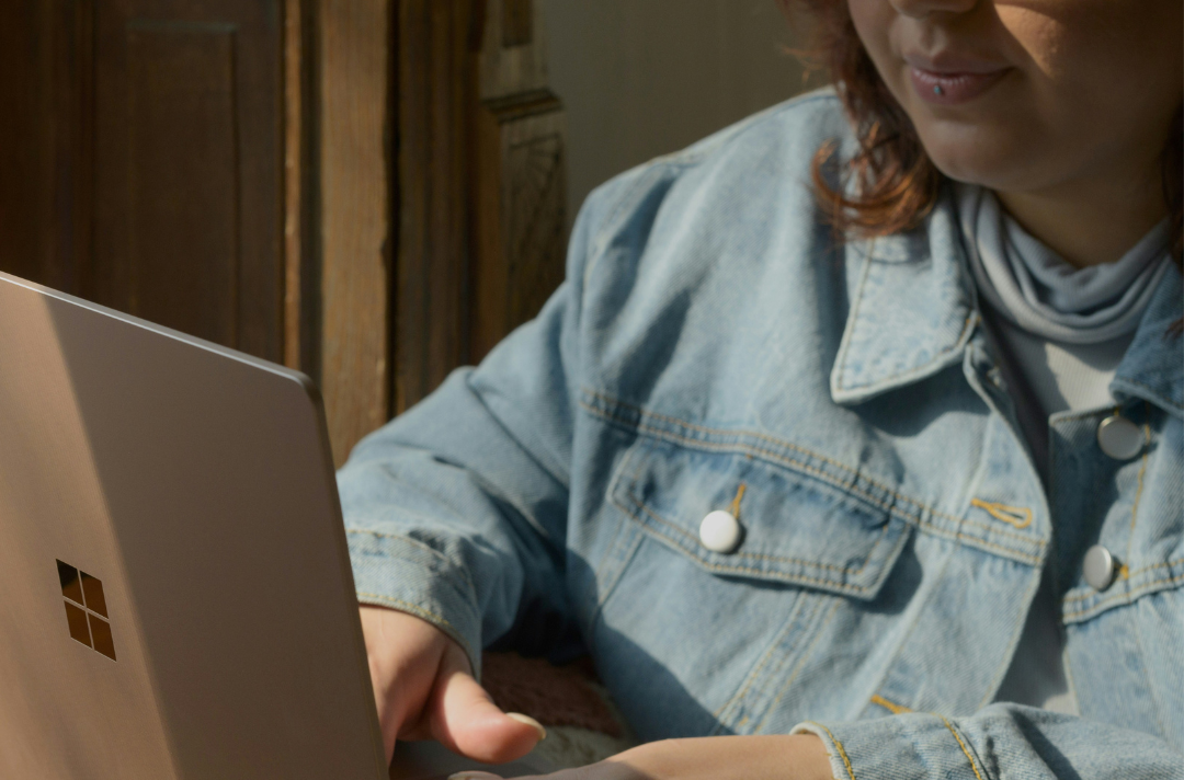 A young woman sitting with a knitted blanket covering her legs and a grey laptop on her lap. Only half of her face is showing in the photograph, and she has dark ginger hair and a lip piercing on her bottom lip. She’s wearing a light blue denim jacket and a light blue roll-neck top. The blanket has yellow, pink, white, and orange patterned squares.