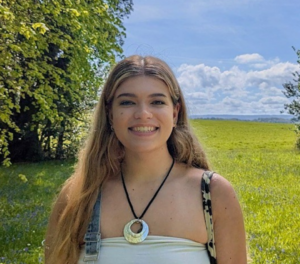 Hannah, an RSBC volunteer. She is standing in a lush green field with trees and grass all around her. The sky is a summery bright blue with just a few white clouds. She is smiling wearing a white tube top, denim dungarees and a necklace with a black neck chain and white circle jewel piece.]