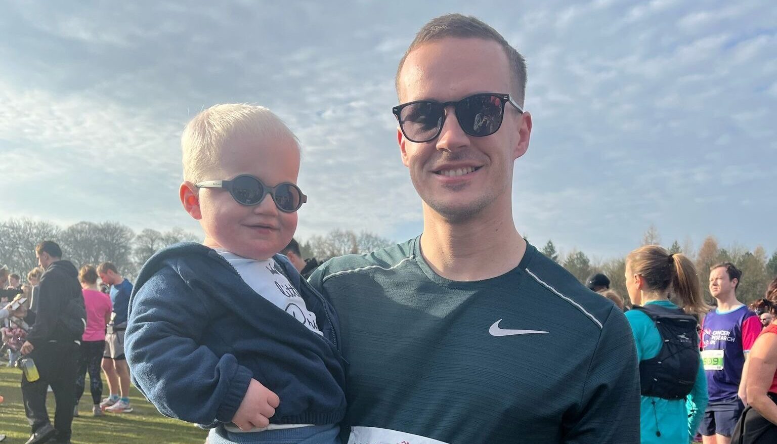 A photograph of Nathan standing in a field holding his son, Alfie with his right arm after completing a running challenge. Nathan is wearing a Nike running vest. Alfie is wearing sunglasses, a white top, hoodie and blue trousers.